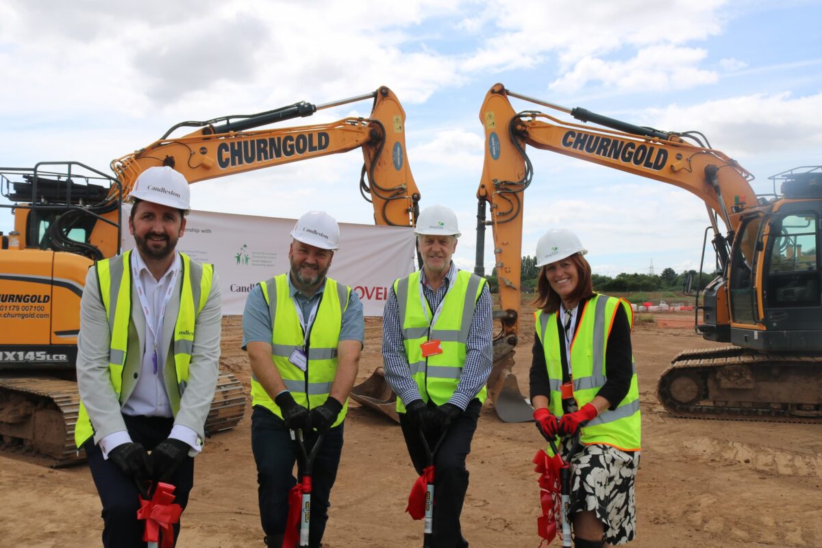 Four people posing with shovels, standing in front of some cranes